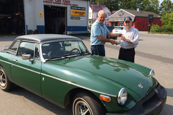 Two men shake hands next to a classic green convertible car in front of an auto repair shop.