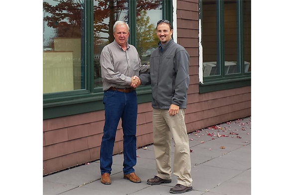 Two men stand outside a building, shaking hands and smiling. One wears a gray jacket and khaki pants, the other a button-down shirt and jeans.